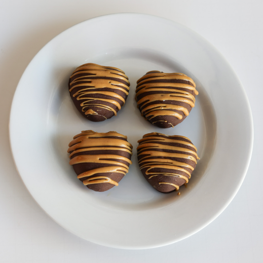 Four heart-shaped chocolate-dipped cheesecake bites with peanut butter drizzle, on a white plate.