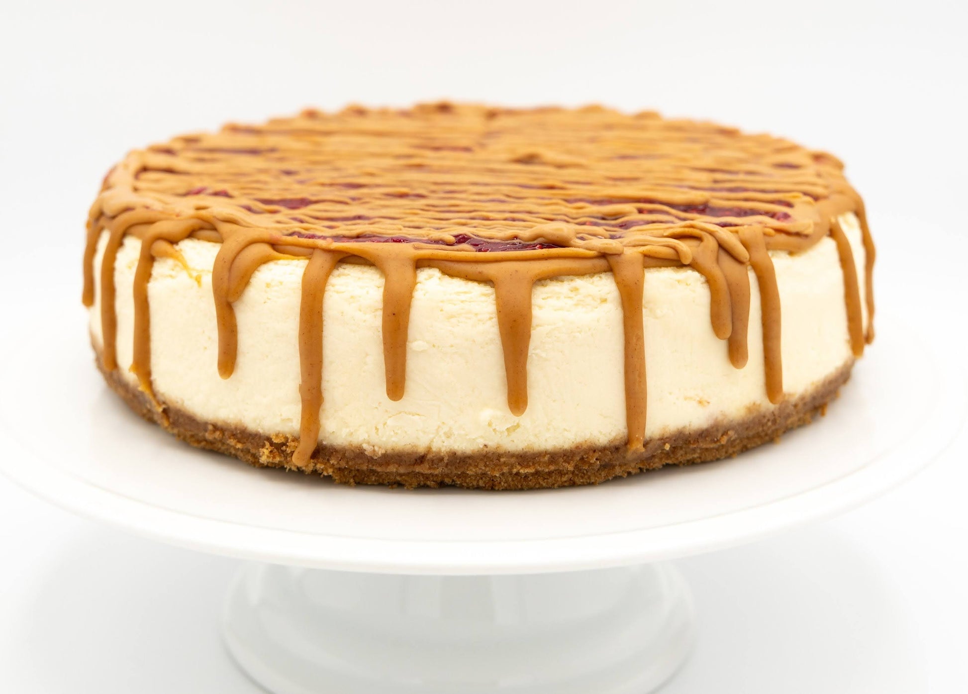 Whole PB & J cheesecake topped with berry sauce and creamy peanut butter drizzle on a white cake stand.
